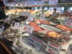 A well-stocked seafood market display featuring a variety of fresh fish and shellfish on ice. Visible species include Alaskan King Salmon, Red Snapper, Black Cod, lobsters, squid, oysters, and octopus. Handwritten signs display prices and product labels. The display is well-organized, with seafood items neatly arranged in rows.