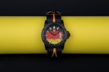 A stylish wristwatch with a bold, colorful dial design is resting on a bright yellow background. The watch face incorporates eye-catching red and yellow patterns, with a clear black bezel and straps that feature the colors black, red, and yellow.