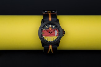 A stylish wristwatch with a bold, colorful dial design is resting on a bright yellow background. The watch face incorporates eye-catching red and yellow patterns, with a clear black bezel and straps that feature the colors black, red, and yellow.