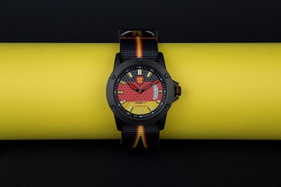 A stylish wristwatch with a bold, colorful dial design is resting on a bright yellow background. The watch face incorporates eye-catching red and yellow patterns, with a clear black bezel and straps that feature the colors black, red, and yellow.