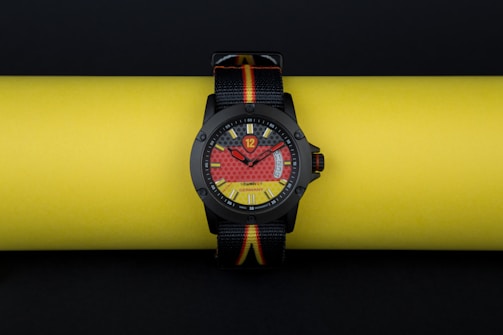 A stylish wristwatch with a bold, colorful dial design is resting on a bright yellow background. The watch face incorporates eye-catching red and yellow patterns, with a clear black bezel and straps that feature the colors black, red, and yellow.