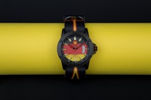 A stylish wristwatch with a bold, colorful dial design is resting on a bright yellow background. The watch face incorporates eye-catching red and yellow patterns, with a clear black bezel and straps that feature the colors black, red, and yellow.