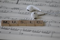 White earphones rest on sheet music with musical notes, accompanied by Scrabble tiles spelling out the word MUSIC.