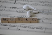 White earphones rest on sheet music with musical notes, accompanied by Scrabble tiles spelling out the word MUSIC.