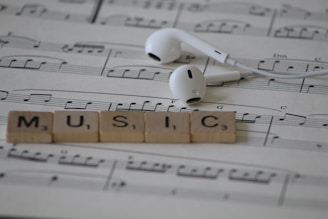 White earphones rest on sheet music with musical notes, accompanied by Scrabble tiles spelling out the word MUSIC.