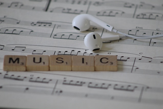 White earphones rest on sheet music with musical notes, accompanied by Scrabble tiles spelling out the word MUSIC.