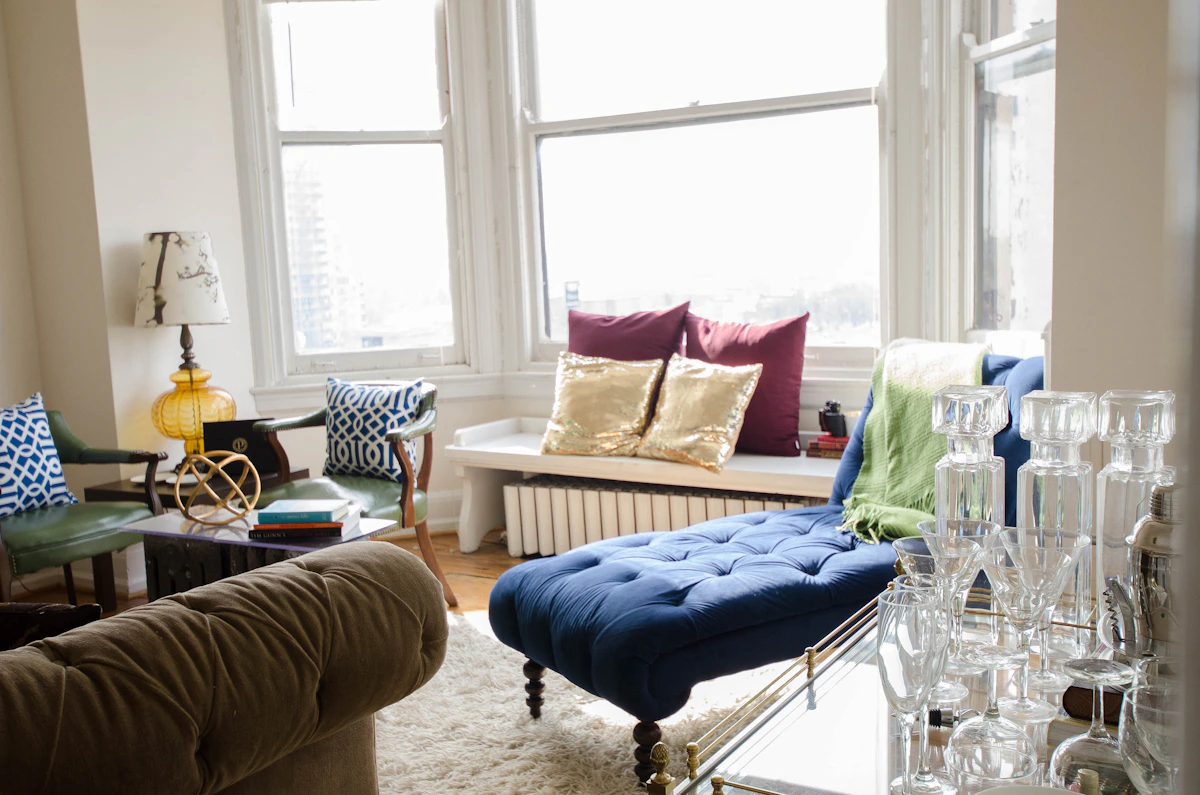 eclectic NYC apartment living room with colorful throw pillows, blue tufted chaise, vintage lamp, and large windows overlooking city