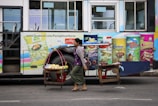 A street vendor carries goods on a yoke past a bus with colorful advertisements for various snacks, including potato chips and crackers.