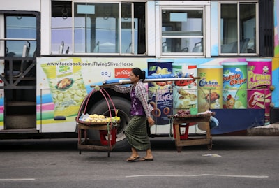 A street vendor carries goods on a yoke past a bus with colorful advertisements for various snacks, including potato chips and crackers.