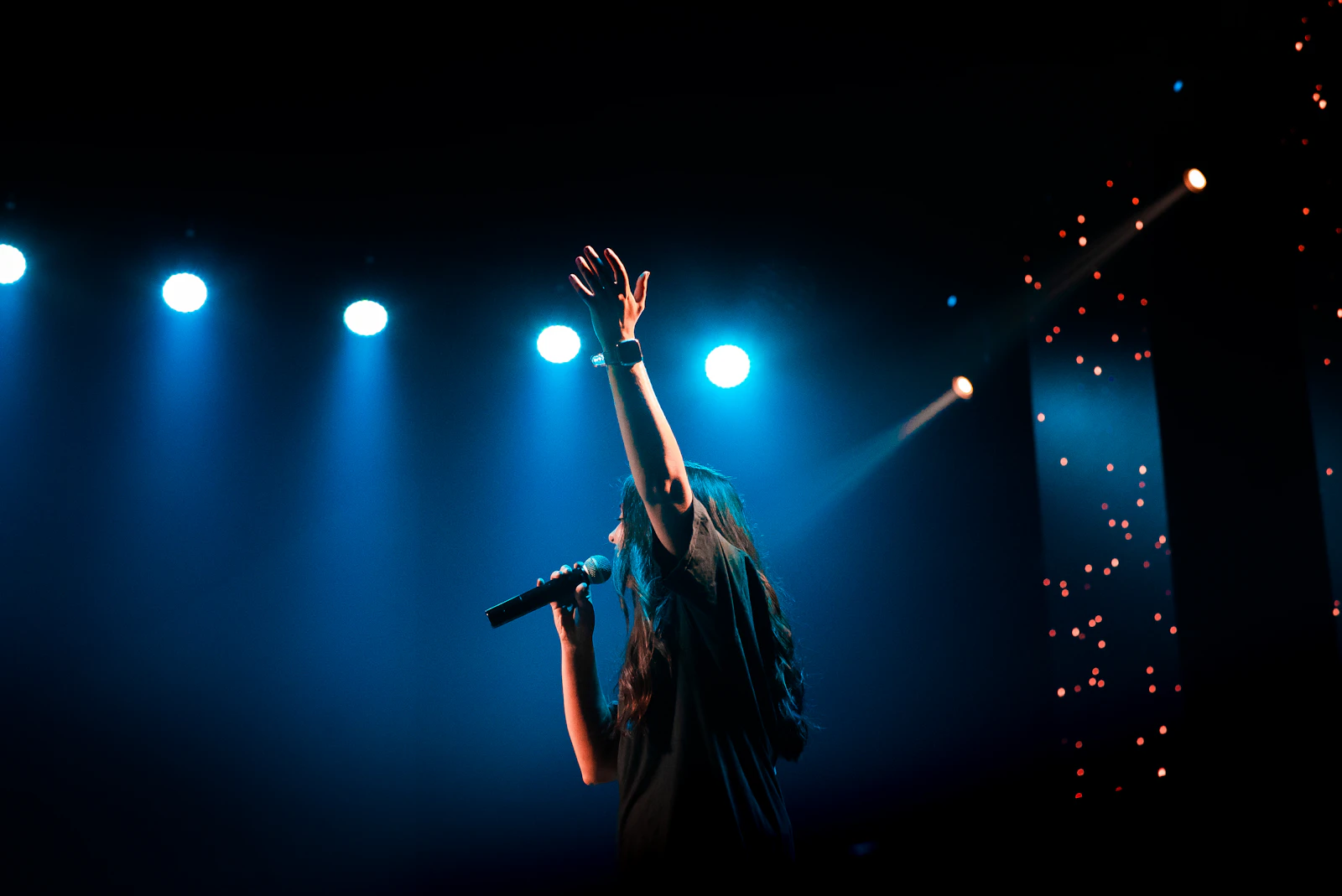 A singer with one arm raised performing on a dark stage under blue spotlights