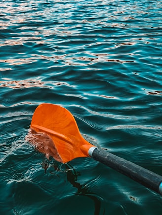 A close-up view of an orange paddle dipping into blue-green water with gentle waves.