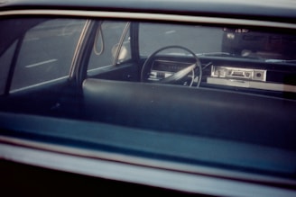 The interior of a vintage car is seen through the rear window, showcasing a classic steering wheel and dashboard. The image conveys a sense of nostalgia, highlighting the simple and robust design typical of older vehicles.