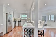 A modern kitchen with wooden flooring, stainless steel appliances, and a marble countertop. The room features white cabinetry and a built-in wine rack at the end of the counter. Barstools with white cushioning are positioned at the counter. The backsplash is composed of small, rectangular tiles in a mosaic pattern. Natural light enters through a wide, rectangular window.