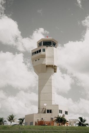 A tall control tower stands prominently against a partly cloudy sky. The structure is rectangular with multiple windows near the top. The base is surrounded by small trees and grass, giving a sense of its location in a natural setting.