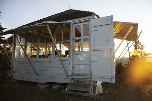 A small, weathered, white wooden cabin with large windows, supported by metal braces, stands amidst a natural setting with greenery. The door is slightly open, and sunlight is streaming inside, casting a warm glow through the windows. Some objects or possibly signs are affixed to the door. The ground leading up to the cabin is gravelly.