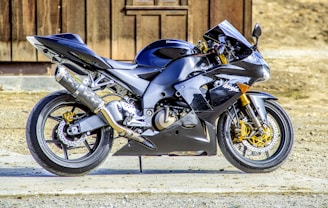 A sleek, modern sport motorcycle with a glossy black finish is parked on a concrete surface. It features distinctive aerodynamic fairings, gold-colored suspension forks, and performance exhaust components. The background consists of a wooden structure and some dry earth.