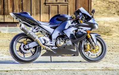 A sleek, modern sport motorcycle with a glossy black finish is parked on a concrete surface. It features distinctive aerodynamic fairings, gold-colored suspension forks, and performance exhaust components. The background consists of a wooden structure and some dry earth.