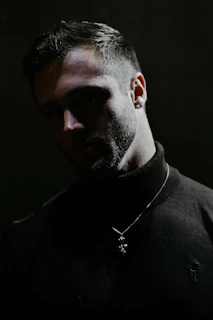 A man with a trimmed beard and short hair is wearing a black turtleneck sweater and a pendant necklace. The lighting is low, casting shadows on his face and creating a mysterious atmosphere.