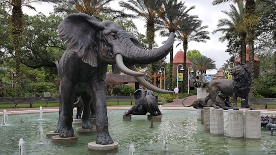 A group of large animal statues, including an elephant and lions, stand in a shallow pool surrounded by palm trees. The setting appears to be a public park or zoo with lush greenery and pathways.