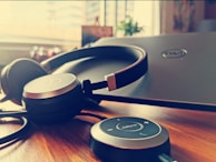 A pair of over-ear headphones rests atop a closed laptop on a wooden table. Beside the headphones is a Jabra control unit, with visible buttons for volume and calls. The background is softly lit by sunlight streaming through a window, with blurred details of a potted plant and urban landscape.