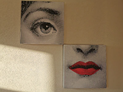 Two separate canvases depict fragments of a face. The upper canvas shows a detailed illustration of an eye, while the lower canvas displays a nose and a pair of red lips. The artworks have a dotted screen effect, reminiscent of pop art. Light from a window casts a shadow on the textured wall beside them.