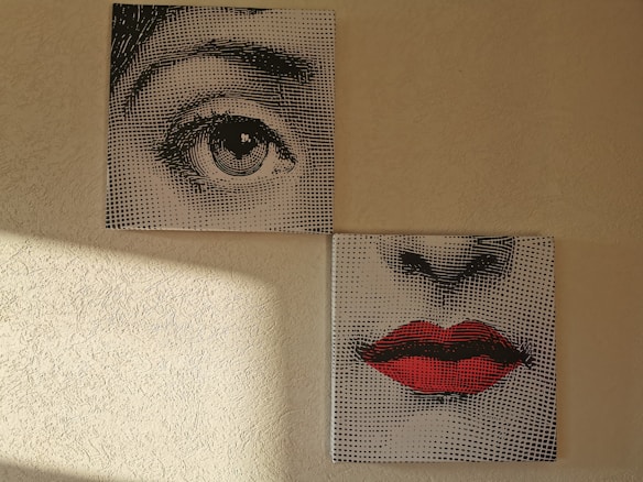 Two separate canvases depict fragments of a face. The upper canvas shows a detailed illustration of an eye, while the lower canvas displays a nose and a pair of red lips. The artworks have a dotted screen effect, reminiscent of pop art. Light from a window casts a shadow on the textured wall beside them.