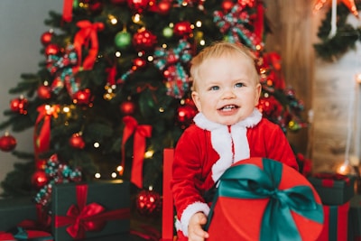 A smiling child in a Santa costume sits in front of a decorated Christmas tree, holding a large red gift with a green ribbon. The tree is adorned with red and green ornaments, ribbons, and lights, creating a festive holiday atmosphere.