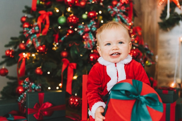 A smiling child in a Santa costume sits in front of a decorated Christmas tree, holding a large red gift with a green ribbon. The tree is adorned with red and green ornaments, ribbons, and lights, creating a festive holiday atmosphere.