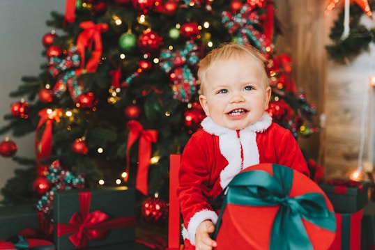 A smiling child in a Santa costume sits in front of a decorated Christmas tree, holding a large red gift with a green ribbon. The tree is adorned with red and green ornaments, ribbons, and lights, creating a festive holiday atmosphere.