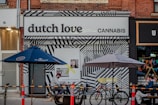 A storefront displays a sign reading 'dutch love cannabis' with a bold, black-and-white geometric patterned facade. Outdoor seating includes tables under blue and grey umbrellas, with people seated and a dog lying on the ground. Bicycles are parked nearby, and a colorful bunting decorates the adjacent store.