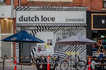 A storefront displays a sign reading 'dutch love cannabis' with a bold, black-and-white geometric patterned facade. Outdoor seating includes tables under blue and grey umbrellas, with people seated and a dog lying on the ground. Bicycles are parked nearby, and a colorful bunting decorates the adjacent store.