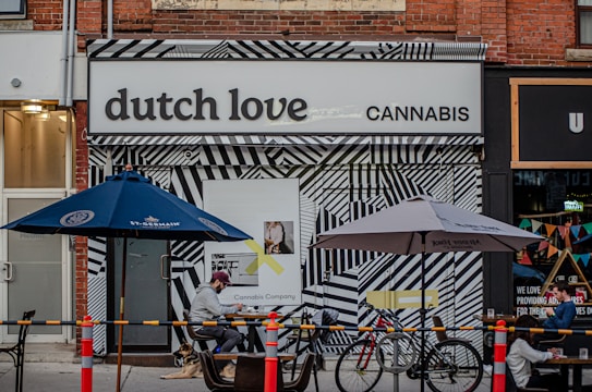 A storefront displays a sign reading 'dutch love cannabis' with a bold, black-and-white geometric patterned facade. Outdoor seating includes tables under blue and grey umbrellas, with people seated and a dog lying on the ground. Bicycles are parked nearby, and a colorful bunting decorates the adjacent store.