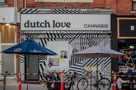 A storefront displays a sign reading 'dutch love cannabis' with a bold, black-and-white geometric patterned facade. Outdoor seating includes tables under blue and grey umbrellas, with people seated and a dog lying on the ground. Bicycles are parked nearby, and a colorful bunting decorates the adjacent store.
