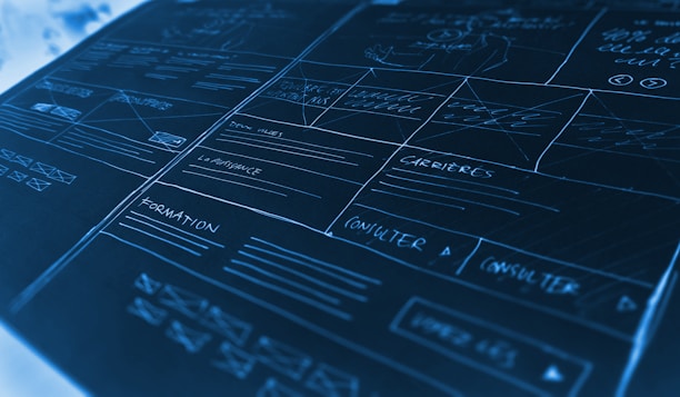 A detailed mock-up or wireframe sketch of a website layout. It features various sections with handwritten notes and placeholders, lines indicating different areas, and labels such as 'carrières' and 'formation'. The design is laid out on a dark, cool-toned surface with white and light blue handwriting, giving a technical and organized appearance. The mock-up includes boxes, lines, and arrows to denote different sections and navigation paths.