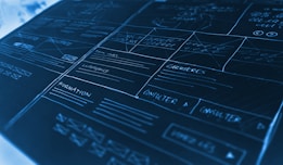 A detailed mock-up or wireframe sketch of a website layout. It features various sections with handwritten notes and placeholders, lines indicating different areas, and labels such as 'carrières' and 'formation'. The design is laid out on a dark, cool-toned surface with white and light blue handwriting, giving a technical and organized appearance. The mock-up includes boxes, lines, and arrows to denote different sections and navigation paths.