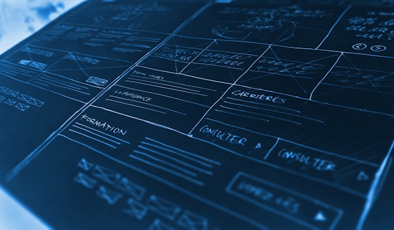 A detailed mock-up or wireframe sketch of a website layout. It features various sections with handwritten notes and placeholders, lines indicating different areas, and labels such as 'carrières' and 'formation'. The design is laid out on a dark, cool-toned surface with white and light blue handwriting, giving a technical and organized appearance. The mock-up includes boxes, lines, and arrows to denote different sections and navigation paths.