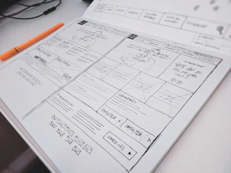 A hand-drawn website wireframe on paper with an orange pen on a desk