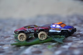 Two toy cars are positioned on a textured surface, surrounded by some green leaves. One car is red with large off-road wheels and a weathered appearance. The other car is a blue and silver pickup truck featuring orange details.