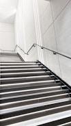 A modern, minimalistic staircase with sleek black railings and bright white lighting integrated into each step. The walls are smooth and white with subtle geometric patterns, creating a clean and futuristic atmosphere.