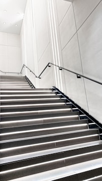 A modern, minimalistic staircase with sleek black railings and bright white lighting integrated into each step. The walls are smooth and white with subtle geometric patterns, creating a clean and futuristic atmosphere.
