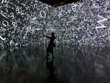 A silhouette of a person stands in front of an immersive digital wall covered in a chaotic array of white letters and symbols on a dark background. The reflective floor adds depth, mirroring the imagery above.
