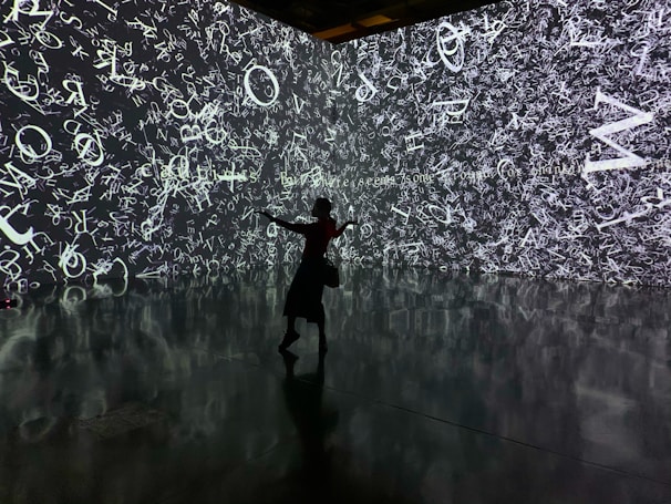 A silhouette of a person stands in front of an immersive digital wall covered in a chaotic array of white letters and symbols on a dark background. The reflective floor adds depth, mirroring the imagery above.