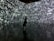 A silhouette of a person stands in front of an immersive digital wall covered in a chaotic array of white letters and symbols on a dark background. The reflective floor adds depth, mirroring the imagery above.