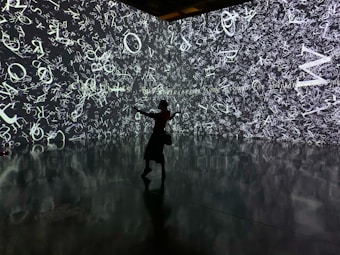 A silhouette of a person stands in front of an immersive digital wall covered in a chaotic array of white letters and symbols on a dark background. The reflective floor adds depth, mirroring the imagery above.