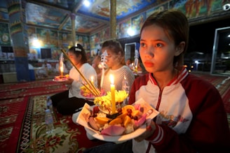 In a vividly decorated interior space with intricate paintings on the walls and ceiling, a group of people are engaged in a ceremonial activity. They are seated on red, ornate carpets, holding trays filled with lit candles, flowers, and incense sticks. The soft, warm candlelight illuminates their faces, creating a serene and contemplative atmosphere.