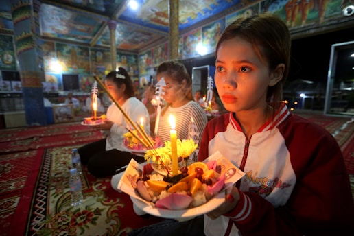 In a vividly decorated interior space with intricate paintings on the walls and ceiling, a group of people are engaged in a ceremonial activity. They are seated on red, ornate carpets, holding trays filled with lit candles, flowers, and incense sticks. The soft, warm candlelight illuminates their faces, creating a serene and contemplative atmosphere.