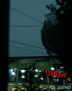 A twilight view of a Trader Joe's storefront with illuminated signs and lights visible through large glass windows. Overhead, electrical wires intersect the sky and silhouetted tree branches frame the scene.