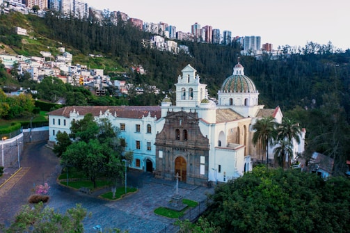A historic church with white walls and a domed roof stands amidst lush greenery. The background features a densely built residential area climbing up a hill. The area is calm with few people visible. Trees surround the building, and palm trees add to the picturesque setting.