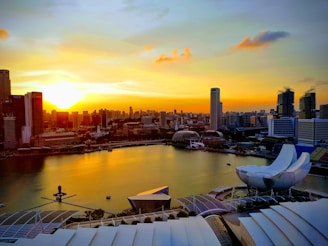 A city skyline during sunset with high-rise buildings and a waterfront. The sun is setting, casting an orange and yellow glow over the scene. The ArtScience Museum with its iconic lotus shape is visible in the foreground. Skyscrapers and other urban structures border the water, creating a modern and dynamic cityscape.
