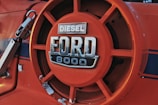 Close-up of an orange Ford 8000 diesel tractor badge featuring a bold, metallic logo with visible signs of wear and some areas of chipped paint.